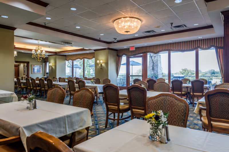 A spacious dining room with multiple tables covered in white tablecloths and surrounded by patterned chairs. The room features large windows with striped valances, allowing natural light to brighten the space. There are chandeliers and wall sconces providing additional lighting, and small flower arrangements are placed on the tables.