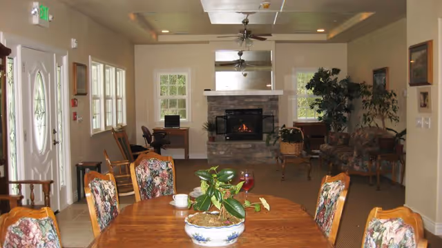 A cozy living room area with a wooden dining table and floral upholstered chairs in the foreground. A potted plant and a glass of red drink are on the table. In the background, there is a stone fireplace with a fire burning, two desks with chairs near windows, a ceiling fan, and a floral patterned sofa with a large potted plant beside it. The room has beige walls and carpeted floor.