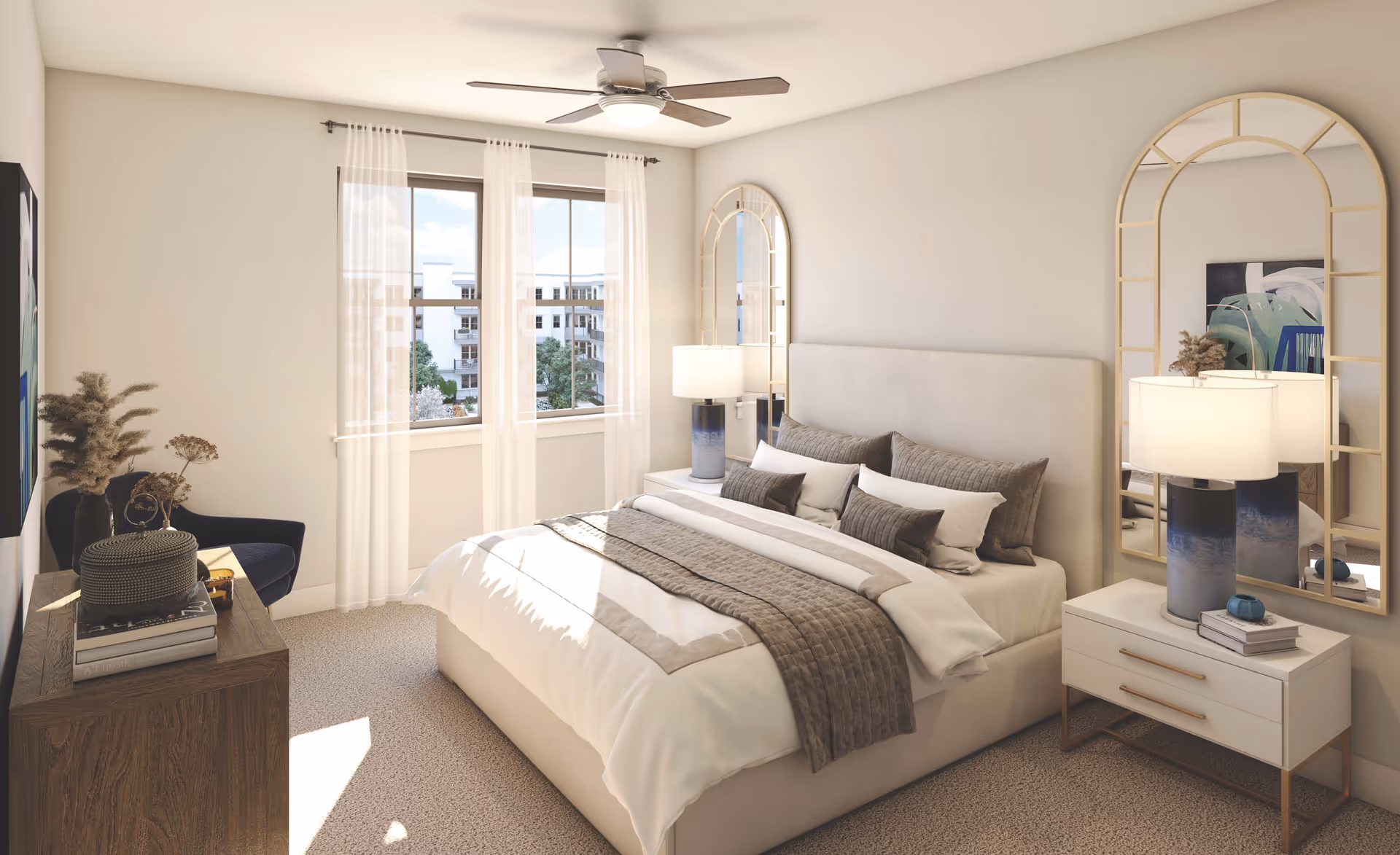 A bright and cozy bedroom with a large bed featuring white and beige bedding and multiple pillows. There are two bedside tables with matching lamps, a large decorative mirror above one bedside table, a ceiling fan, a window with sheer white curtains letting in natural light, a wooden dresser with books and decorative items, and a dark blue armchair in the corner.