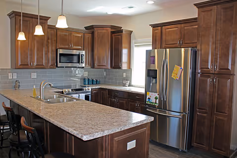 Modern kitchen with dark wooden cabinets, a large granite countertop island with a double sink, stainless steel refrigerator with magnets, microwave, and stove. There are three pendant lights hanging above the island and three bar stools along the island.