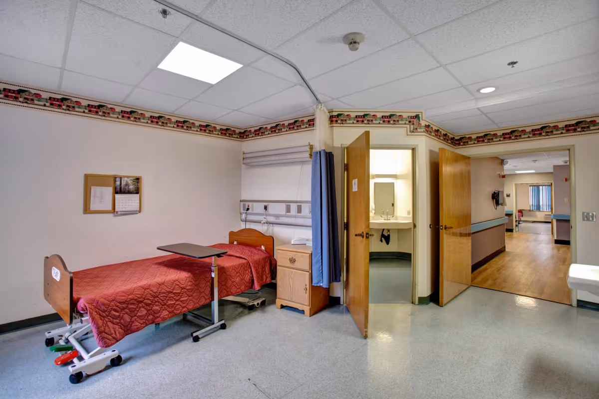 A hospital-style room with a single bed covered in a red quilt, an overbed table, a small wooden nightstand, and a partially open door leading to a small sink area. The room has a ceiling-mounted curtain track with a blue privacy curtain, light-colored walls with a decorative border near the ceiling, and a hallway visible through another open door.