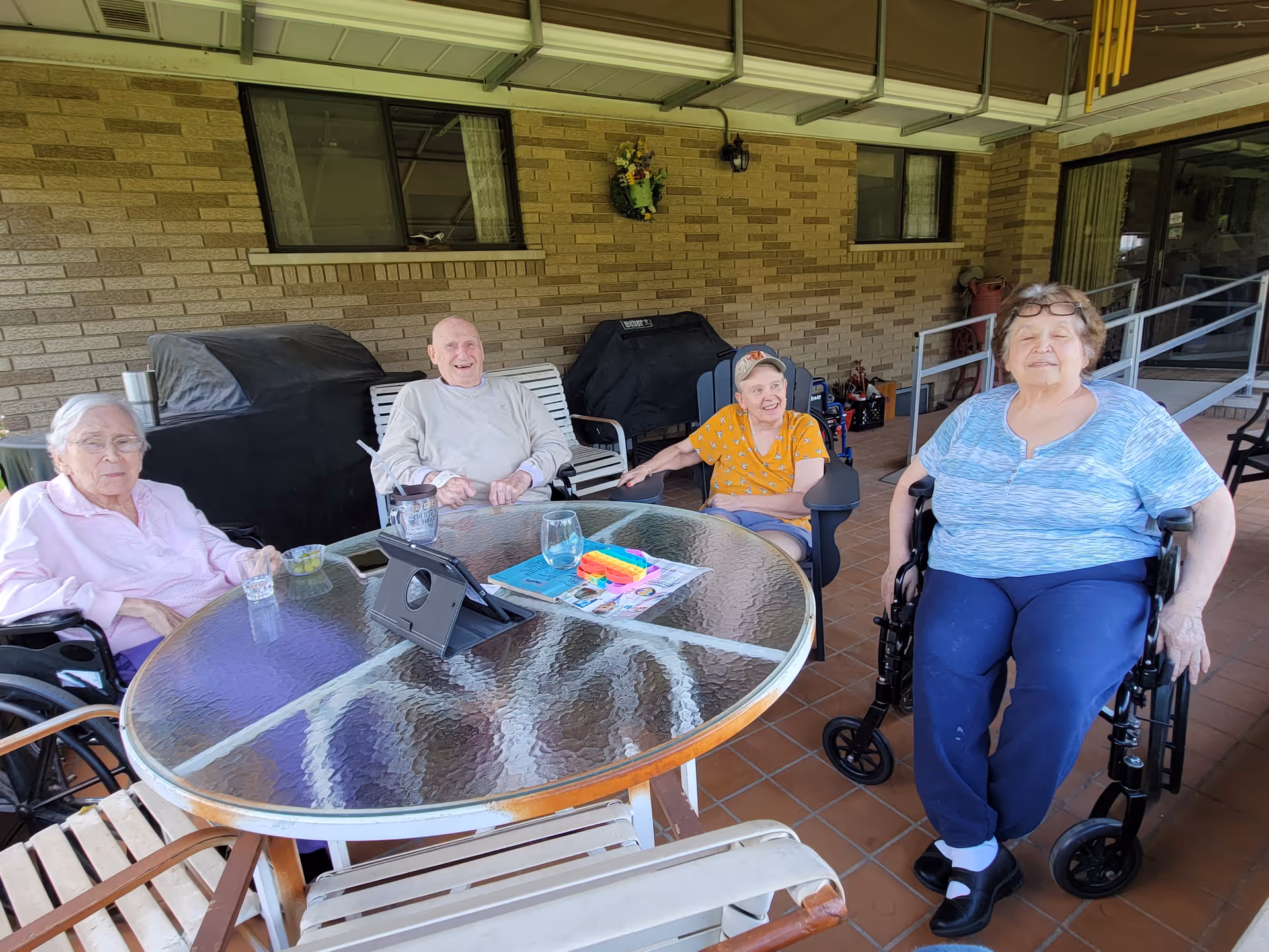 Four elderly individuals sitting around a glass outdoor table on a covered patio. Two are in wheelchairs, one is seated on a bench, and one is in a chair. The table has a tablet, a glass of water, and some colorful items on it. The background shows a brick wall with two windows and a covered grill.