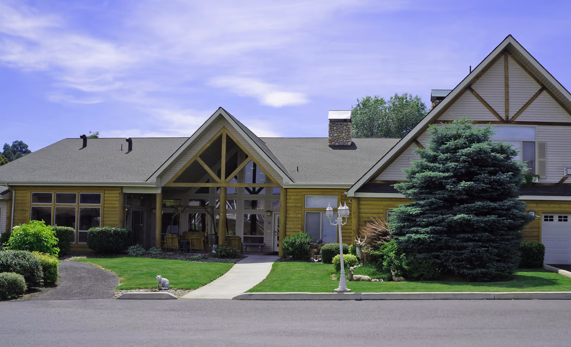 Front exterior view of a single-story building with a peaked roof and large windows. The building is surrounded by well-maintained green lawns, bushes, and a large evergreen tree. A paved walkway leads to the entrance, and a decorative street lamp is visible on the lawn.