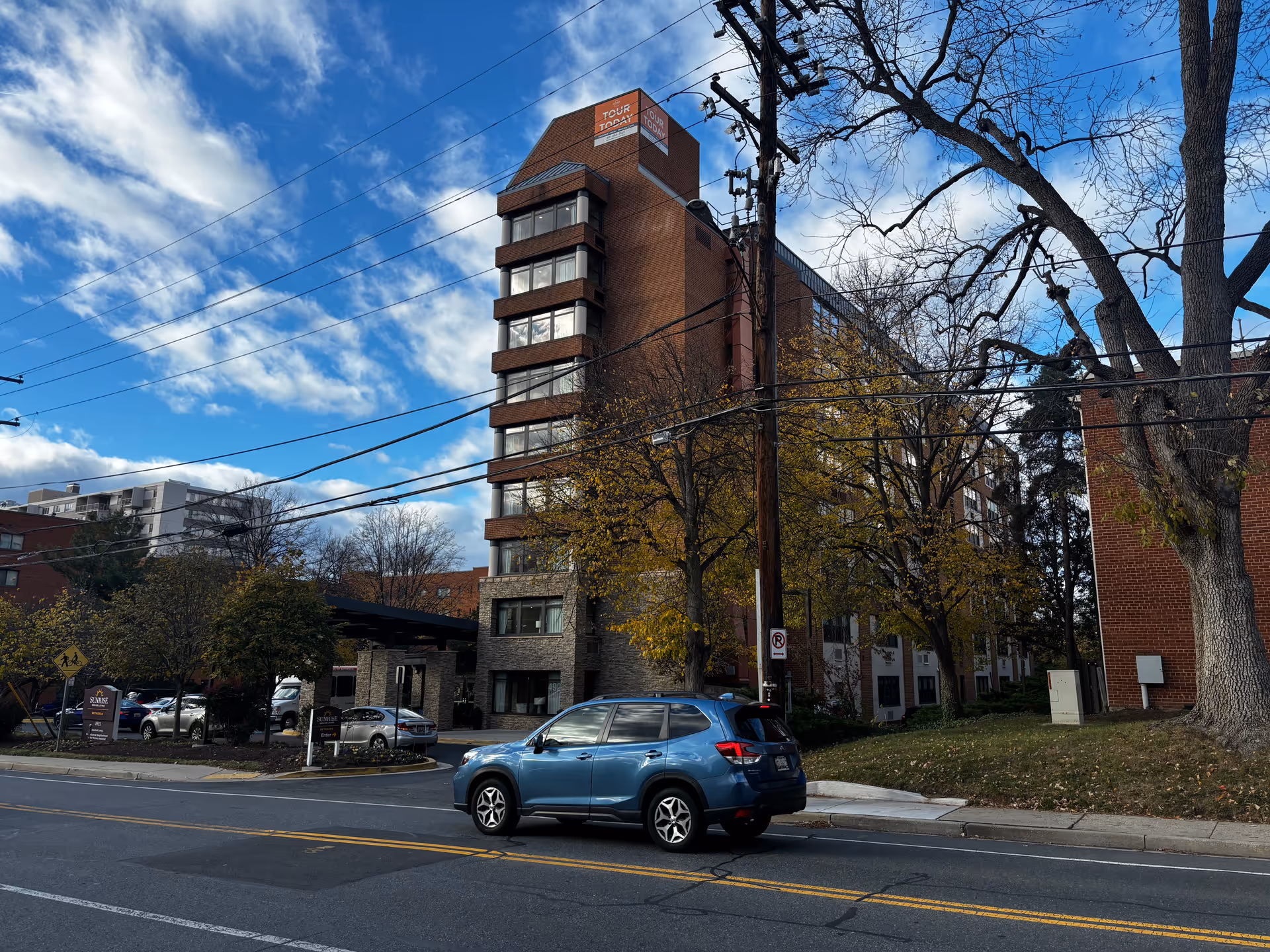 Exterior view of a multi-story brick building with a sign for Sunrise of Bethesda at the entrance. A blue SUV is parked on the street in front of the building. Trees with autumn leaves and a partly cloudy blue sky are visible.