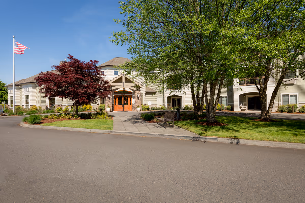 Front exterior view of Brookdale Vancouver Orchards facility with a paved driveway, green lawn, trees, a bench, and an American flag on a flagpole under a clear blue sky.