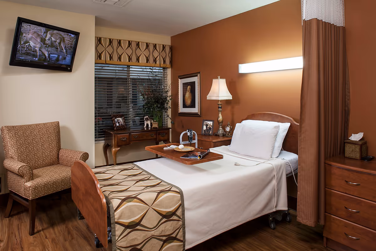 A cozy bedroom in a senior living facility featuring a single bed with white linens and a patterned bed runner, a wooden bedside table with a lamp and framed photos, a patterned armchair, a wall-mounted TV showing a nature scene, a wooden desk with decorative items, and a window with blinds and a valance.