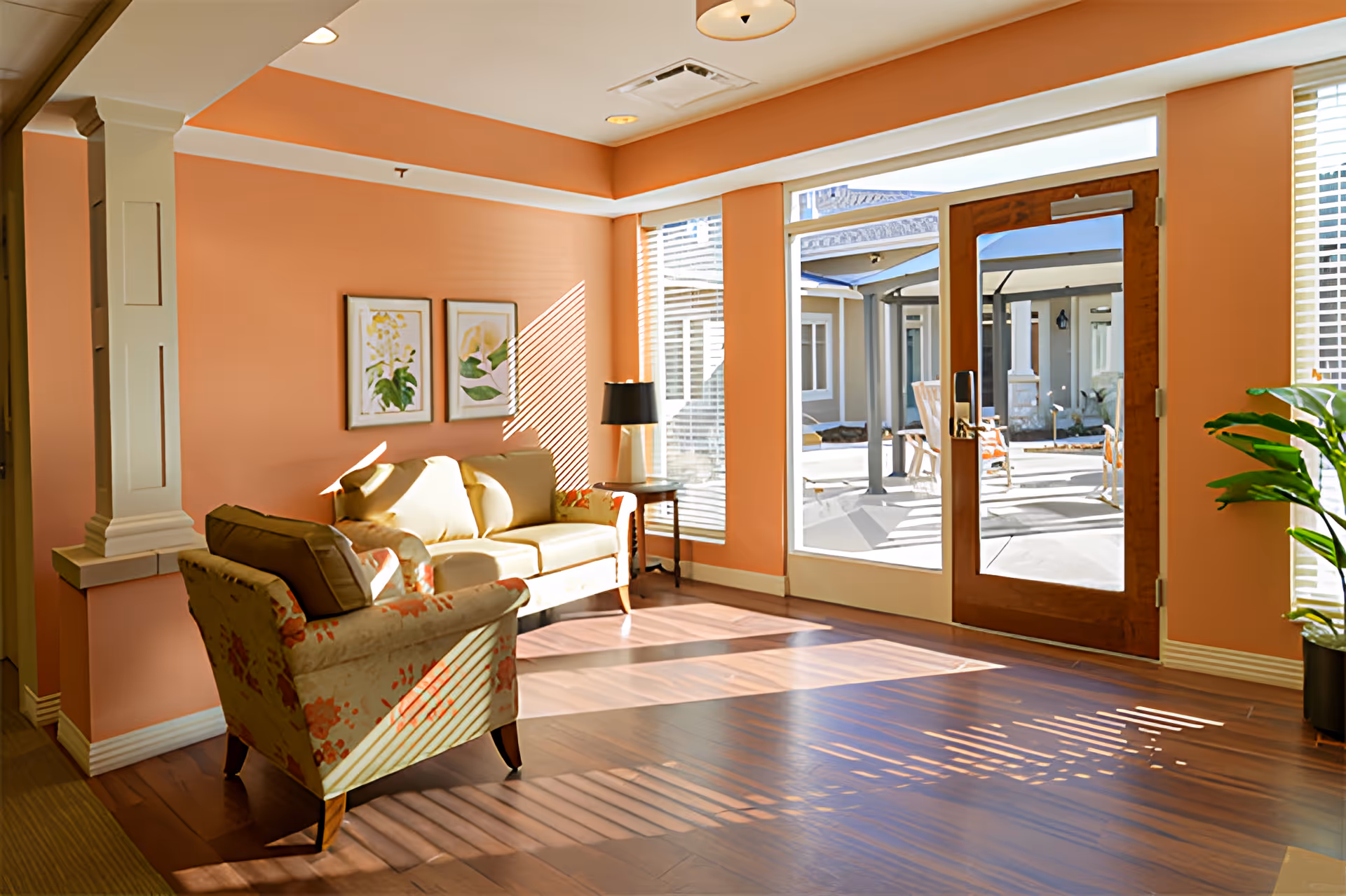 Sunlit seating area with a sofa and armchair on wood floors and a glass door opening to an outdoor courtyard.