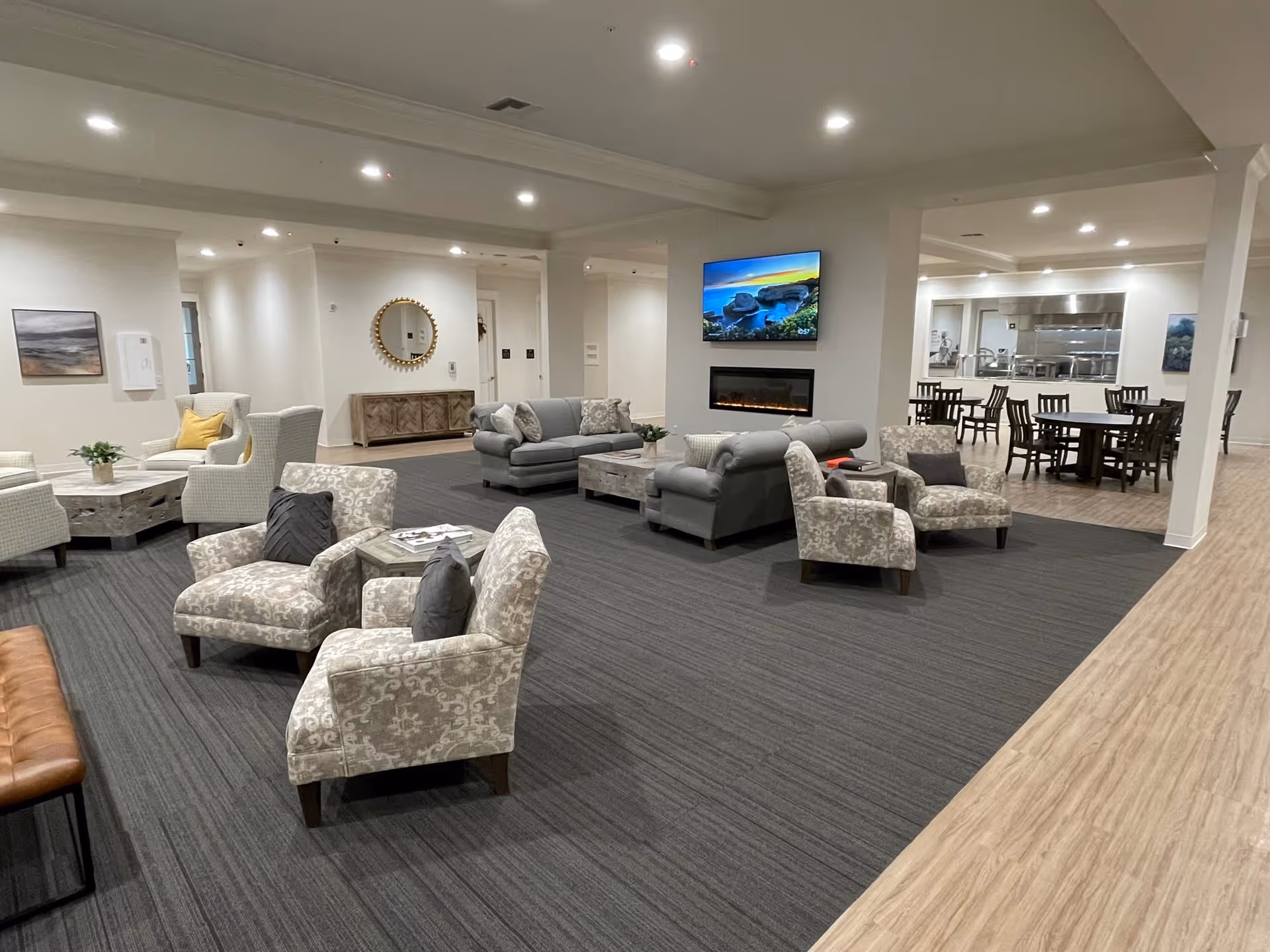 A spacious and well-lit common area in a senior living facility featuring multiple seating arrangements with patterned armchairs, sofas, and coffee tables. A modern electric fireplace is mounted on a central wall beneath a flat-screen TV displaying a scenic image. In the background, there is a dining area with several wooden tables and chairs, and a kitchen area with stainless steel appliances and counters. The flooring transitions from carpet in the seating area to wood-like flooring in the dining and kitchen areas.