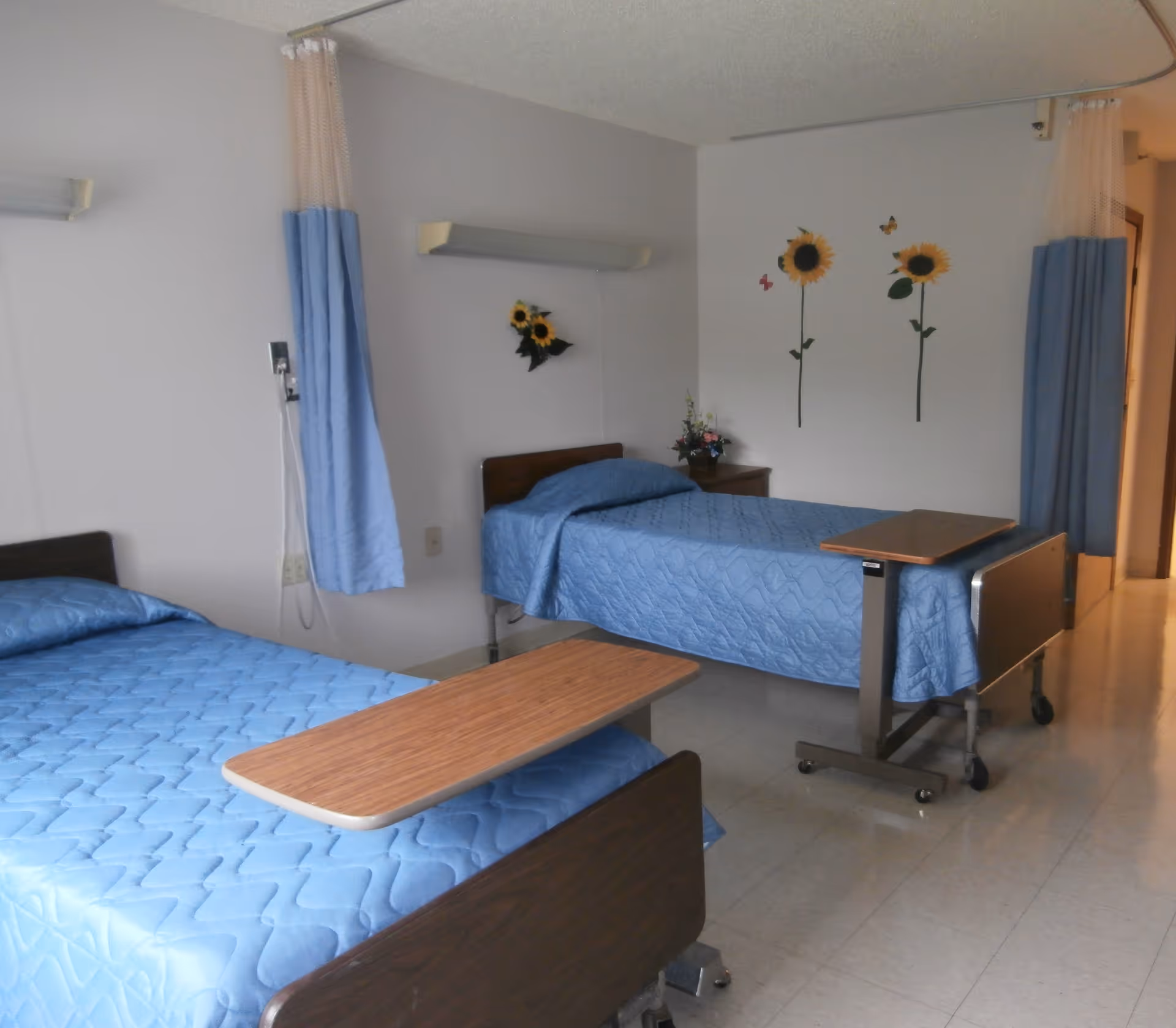 A room with two hospital-style beds covered with blue quilts, each equipped with an overbed table. The room has light-colored walls decorated with sunflower wall decals and a small floral arrangement on a bedside table. Blue privacy curtains hang beside each bed.