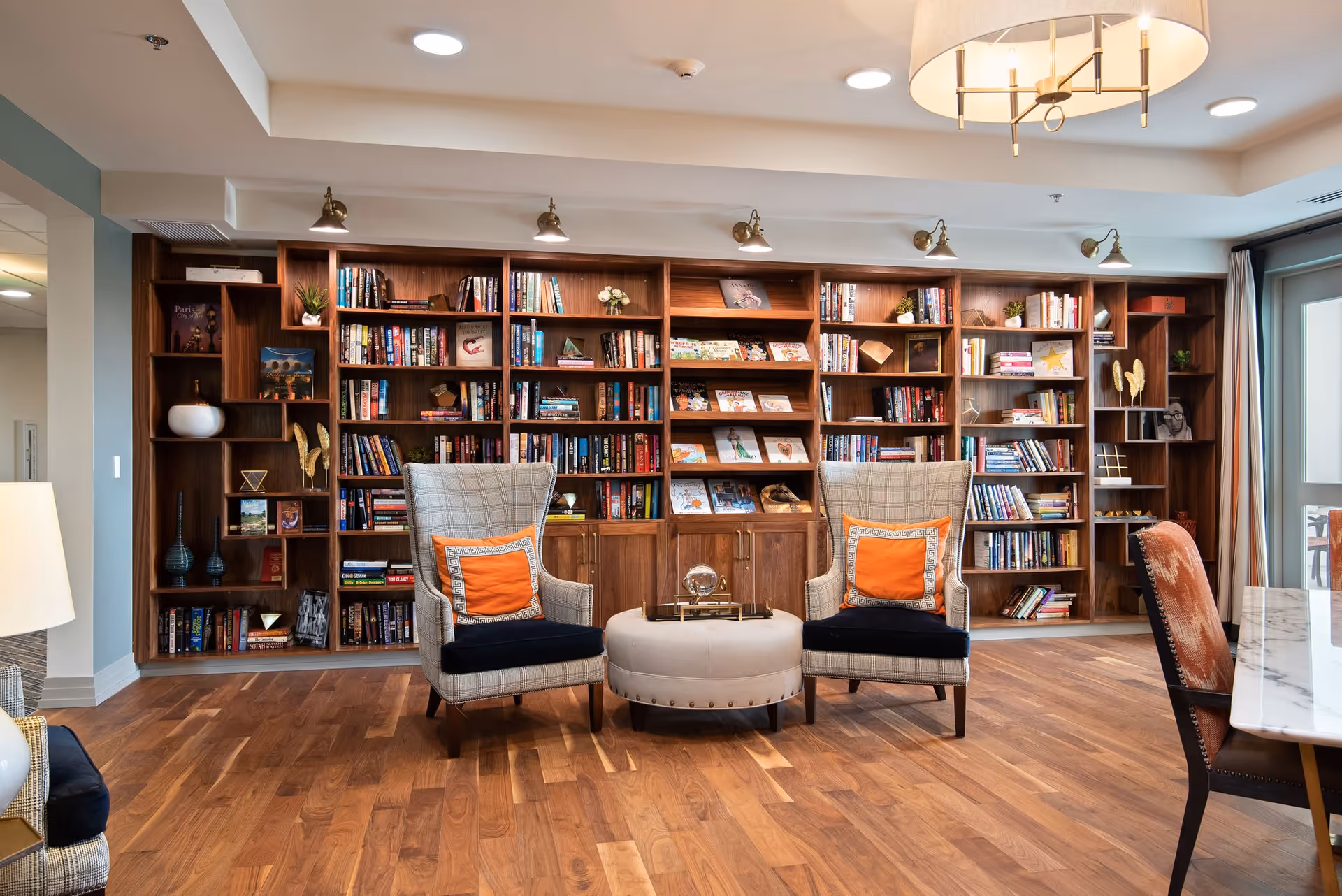 A cozy library or reading area in a senior living facility featuring a large wooden bookshelf filled with books and decorative items. In front of the bookshelf are two cushioned armchairs with orange pillows and a round ottoman between them. The room has wooden flooring, a modern ceiling light fixture, and a table with chairs partially visible on the right side.