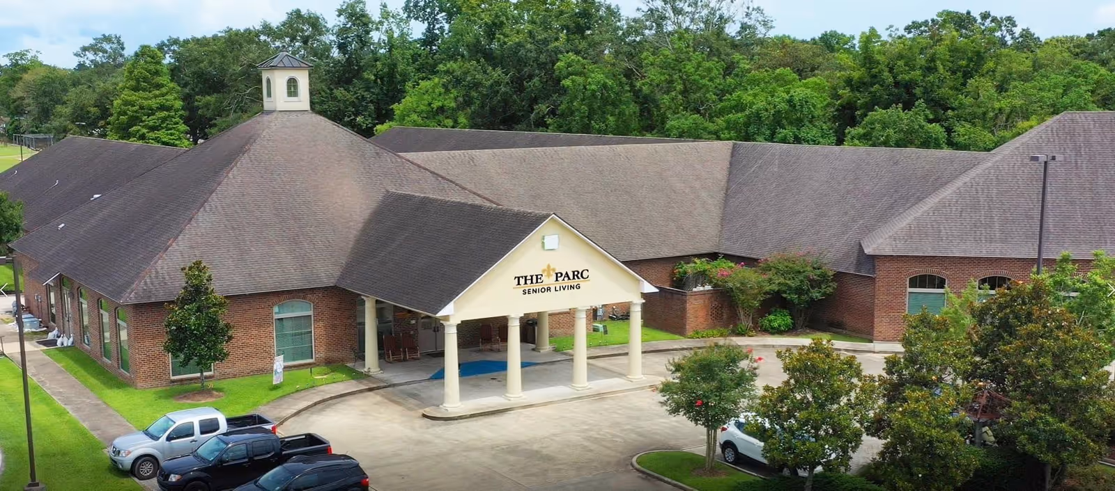 Front exterior of The Parc Senior Living building with a covered entrance, circular driveway, parked cars, and surrounding trees.
