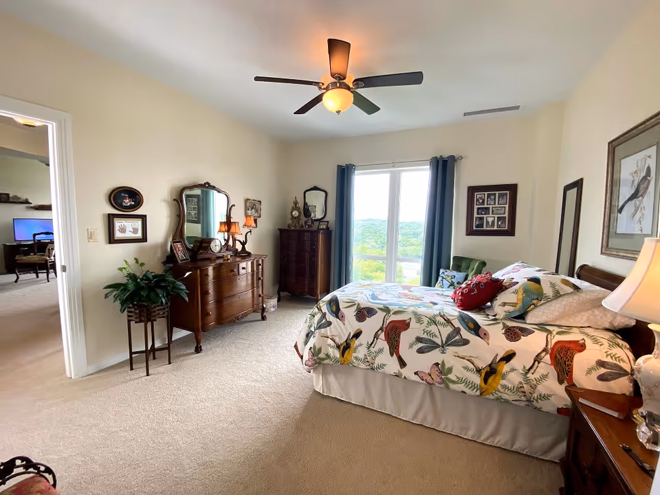 A bright bedroom with a large window covered by blue curtains, a bed with colorful bird-patterned bedding, a wooden dresser with a mirror, a tall chest of drawers, framed pictures on the walls, a ceiling fan with a light, and a potted plant near the doorway leading to another room.