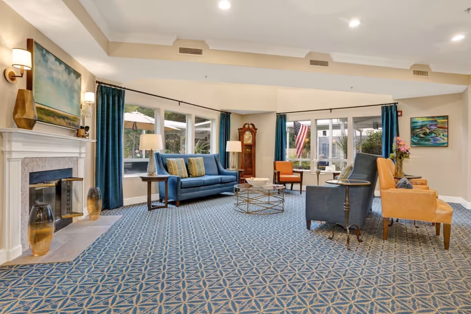 A bright communal lounge with sofas and armchairs arranged around a coffee table, a fireplace, grandfather clock and large windows.