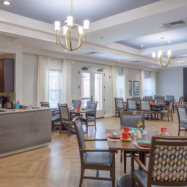 A bright and spacious dining room with multiple wooden tables and chairs arranged neatly. Each table is set with red cups, green napkins, and plates, with some tables featuring small flower centerpieces. The room has large windows with white curtains, two modern chandeliers hanging from the ceiling, and a kitchen counter visible on the left side.