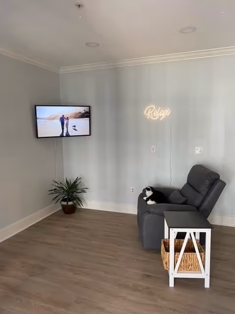 A cozy living area with a gray recliner chair, a small white side table with a wicker basket underneath, a potted plant on the floor, a wall-mounted TV displaying a couple walking on a beach, and a neon sign on the wall that says 'Relax'.