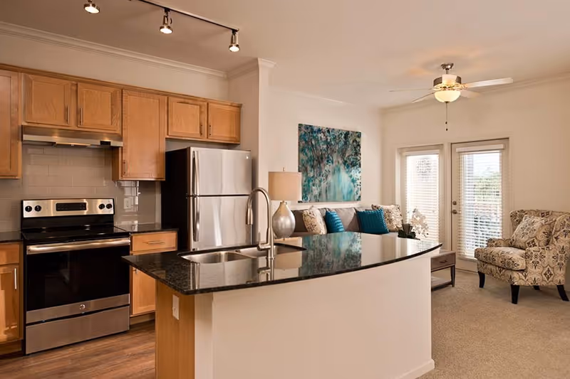 Interior view of a modern kitchen and living room area in a senior living facility. The kitchen features wooden cabinets, a stainless steel refrigerator and oven, a black granite countertop island with a sink and faucet. The living room has a patterned armchair, a sofa with blue and patterned cushions, a table lamp, a coffee table, and a large abstract painting on the wall. There are glass doors with blinds leading outside.