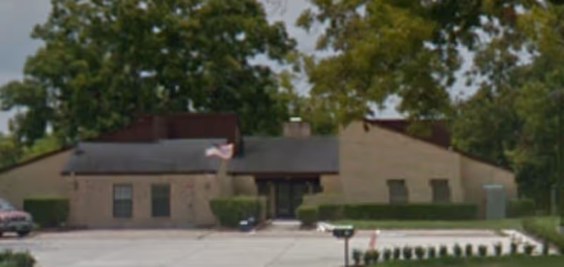 Exterior front view of a single-story senior living facility building with a sloped roof, beige brick walls, several windows, and an American flag near the entrance. There are bushes along the front of the building and a parking lot in the foreground.