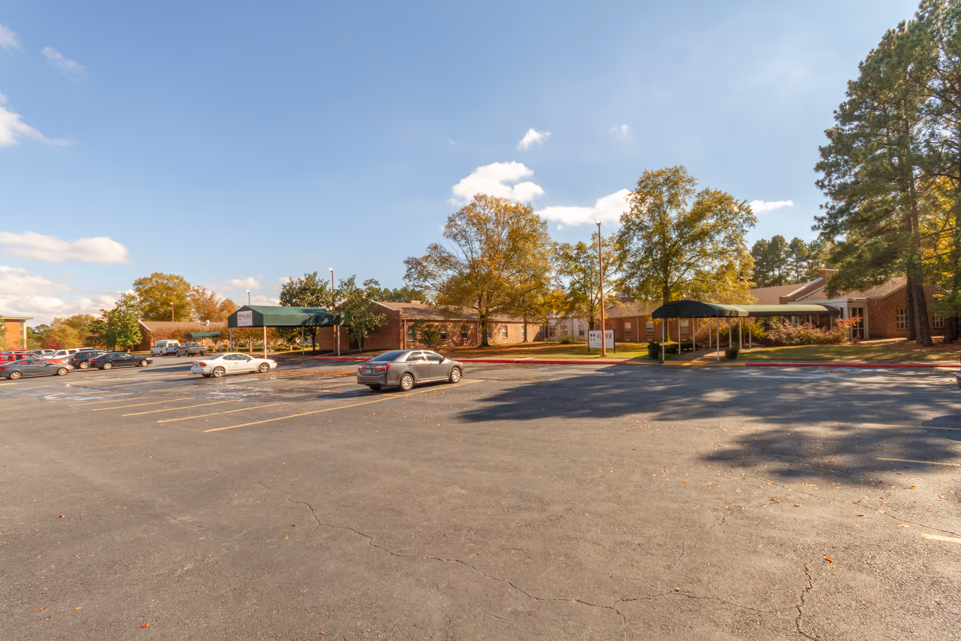 Parking lot and entrance canopies in front of a low brick nursing facility surrounded by trees under a blue sky.
