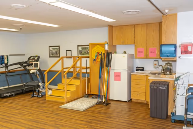 Interior therapy/exercise room with a treadmill, wooden rehabilitation steps, and a small kitchenette with a refrigerator.