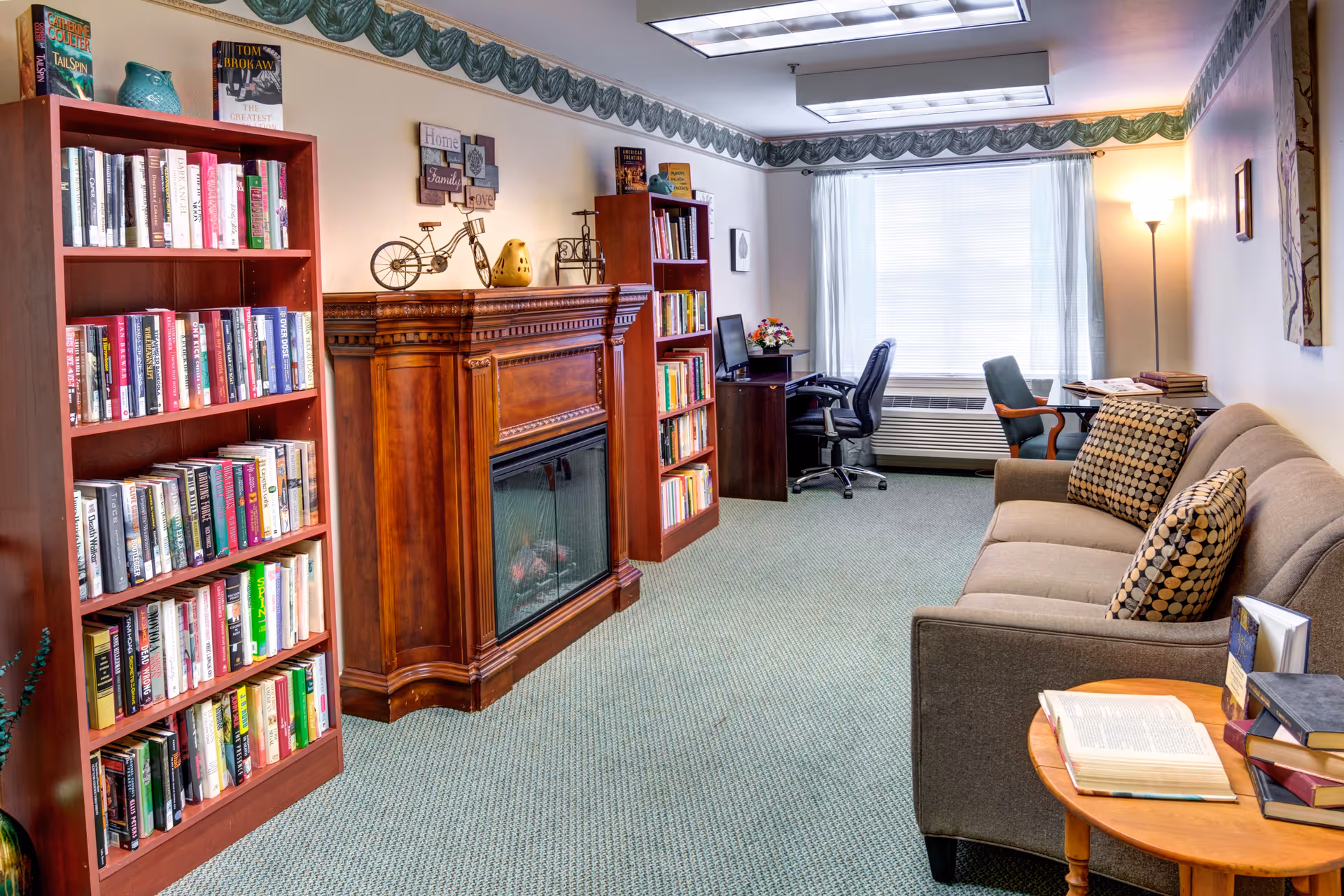 A cozy living room with a beige sofa adorned with patterned cushions, a wooden coffee table with open books, two tall wooden bookshelves filled with books, a decorative wooden fireplace, a desk with a computer and office chair near a window with white curtains, and a floor lamp in the corner.