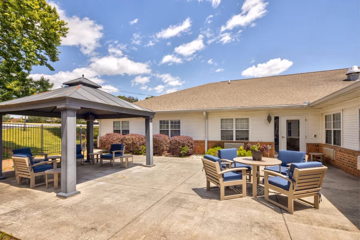 Outdoor patio area at TerraBella Marchbanks featuring a covered gazebo with seating and a round table with chairs on a concrete surface, surrounded by a white building with brick accents, windows, and a door. The sky is partly cloudy and there is greenery including bushes and a tree nearby.