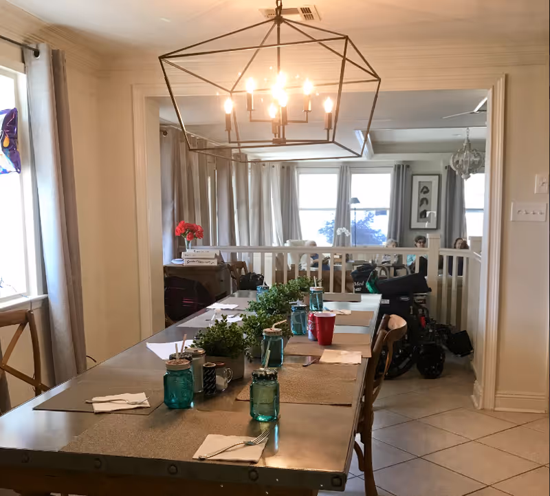 Dining room with a long table set with mason jars, placemats and greenery under a geometric chandelier, opening onto a bright living area.