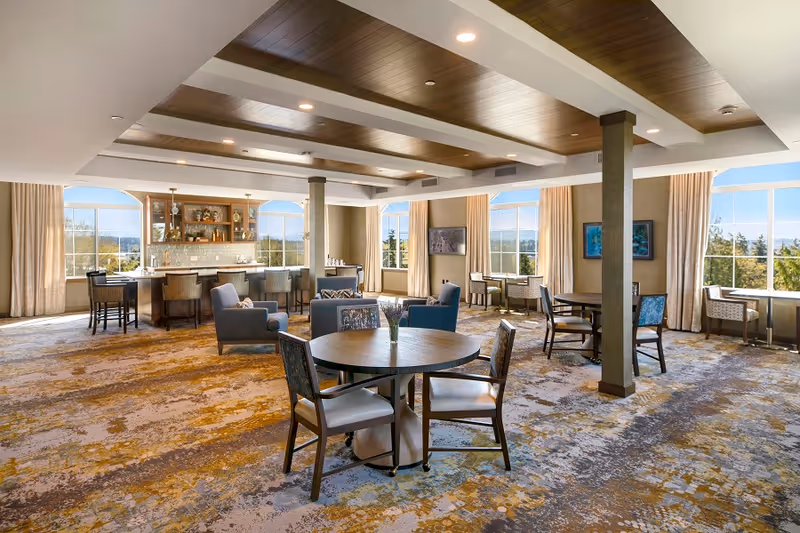 Spacious and well-lit common area with multiple round tables and chairs, comfortable armchairs, large windows with beige curtains letting in natural light, a bar area with stools, and a ceiling with wooden panels and recessed lighting.