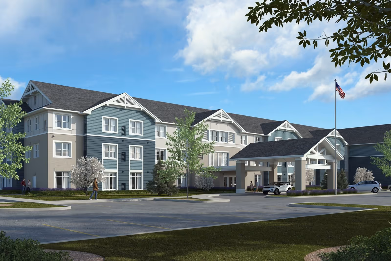 Exterior view of a three-story senior living facility building with a covered entrance and an American flag on a flagpole. There are a few people walking near the building and cars parked in front. The sky is blue with some clouds and there are trees and landscaping around the building.