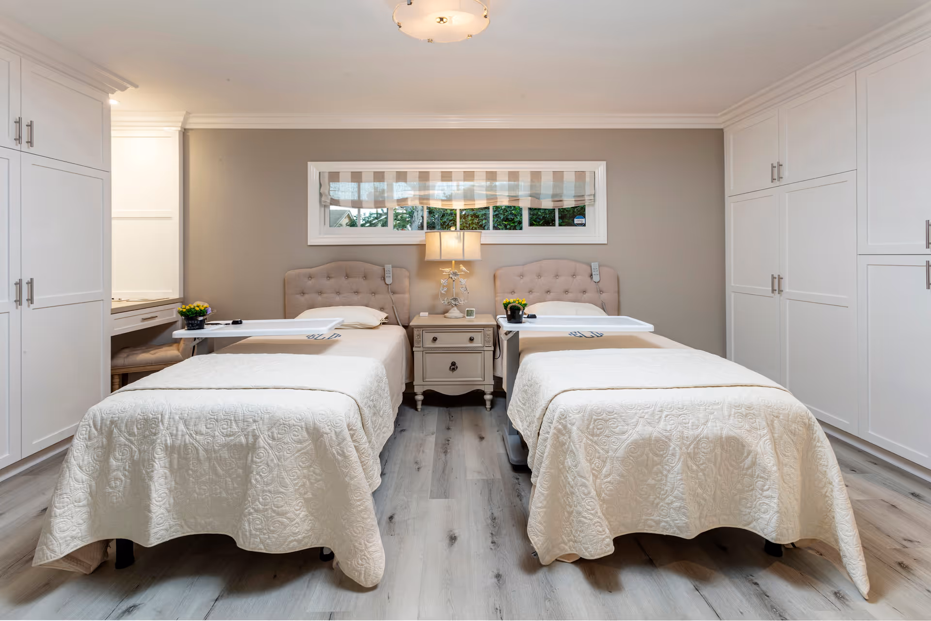 A clean and well-lit bedroom with two single beds side by side, each with a beige quilted bedspread and a tufted headboard. Between the beds is a small nightstand with a decorative lamp and a small clock. Above the nightstand is a rectangular window with a striped Roman shade. The room has light-colored wooden flooring and white built-in cabinets on both sides.