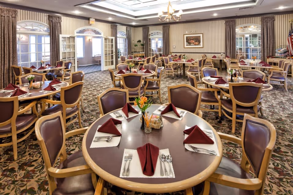 A spacious dining room with multiple round tables set with maroon folded napkins, silverware, and small flower arrangements. The room features patterned carpet, striped wallpaper, large windows with curtains, and a chandelier hanging from the ceiling.