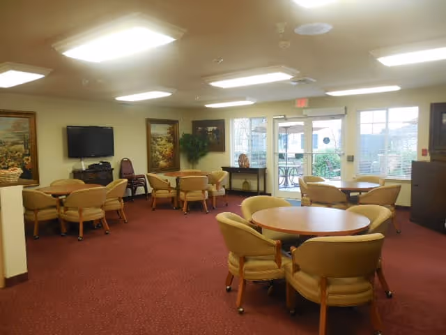A senior living community common area with multiple round tables surrounded by cushioned chairs on a red carpet. The room has large windows and a glass door leading outside, a flat-screen TV mounted on the wall, framed paintings, and potted plants.