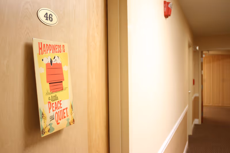 A hallway in an assisted living facility with beige walls and carpeted floor. A wooden door labeled with the number 46 has a decorative sign featuring Snoopy lying on his red doghouse with the text 'Happiness is a little peace and quiet.'