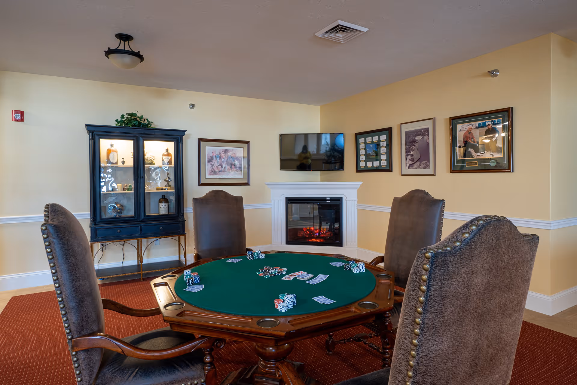 A cozy room with a round poker table covered in green felt, surrounded by four upholstered chairs with wooden arms. The table has poker chips and playing cards on it. In the background, there is a white electric fireplace with a flat-screen TV mounted above it. The walls are decorated with framed pictures and a glass display cabinet with various items inside. The room has beige walls, a red carpet, and a ceiling light fixture.