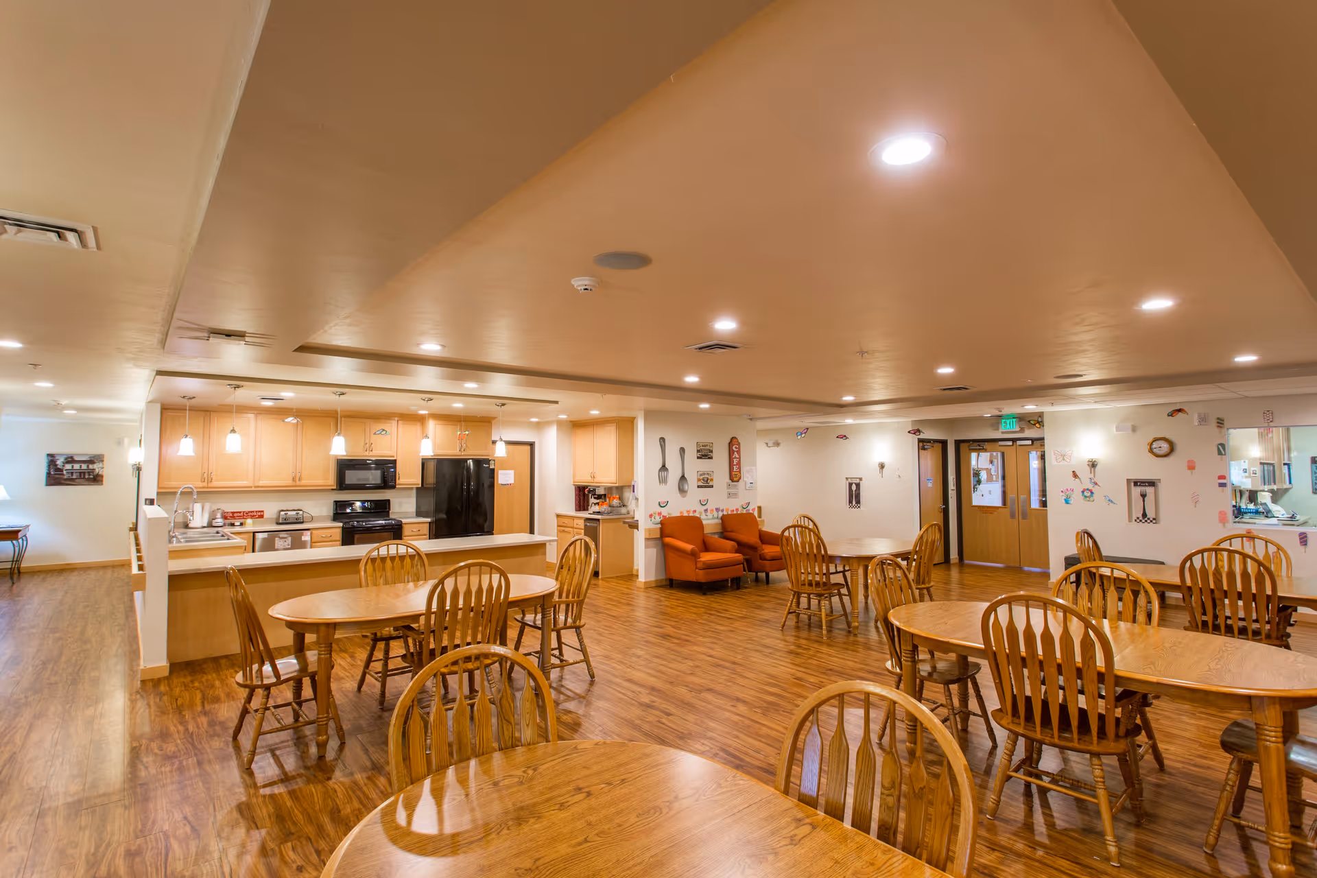 Open communal dining area with wooden tables and chairs and a kitchen counter in a senior living facility.