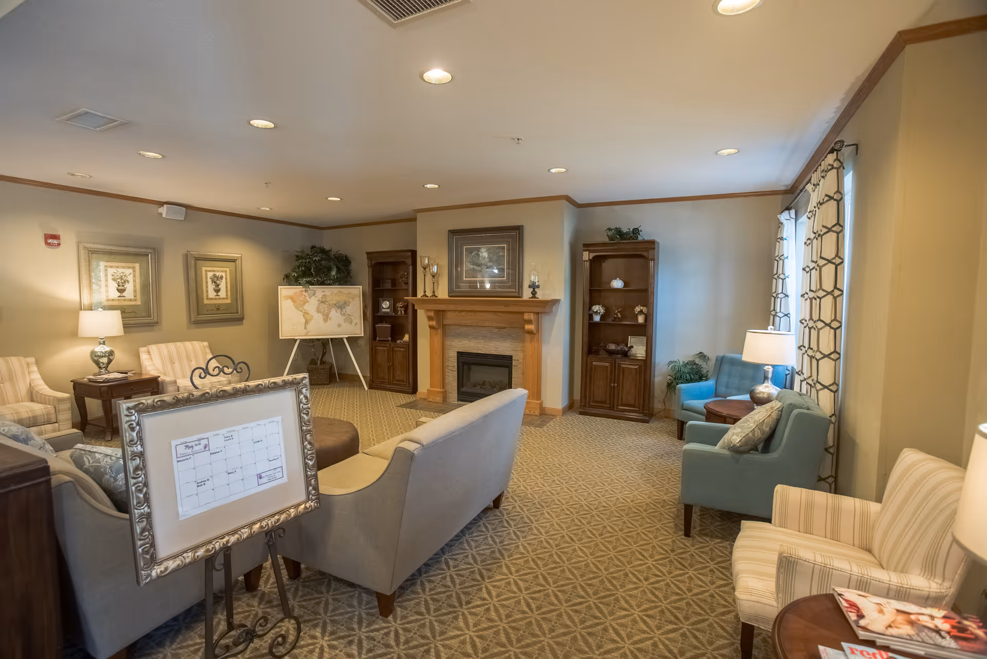 A cozy living room area in a senior living facility with multiple upholstered chairs and sofas arranged around a central fireplace. The room features a patterned carpet, framed artwork on the walls, a world map on an easel, wooden bookshelves with decorative items, and several table lamps providing warm lighting. Large windows with patterned curtains allow natural light into the space.