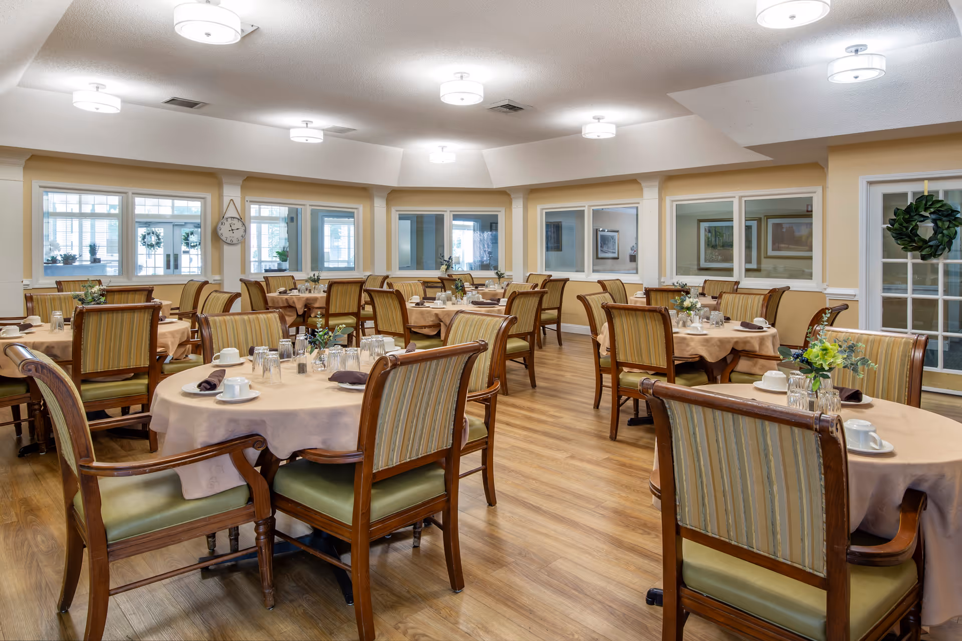Bright, spacious dining room with round tables set for meals and wooden chairs in a senior living facility.