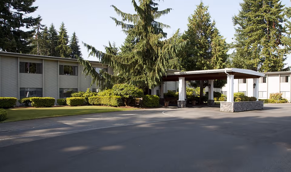 Exterior view of a senior living facility with a covered entrance, surrounded by green bushes and tall evergreen trees under a clear sky.