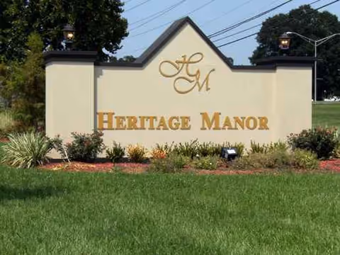 A beige Heritage Manor sign with gold lettering, flanked by shrubs and a green lawn.