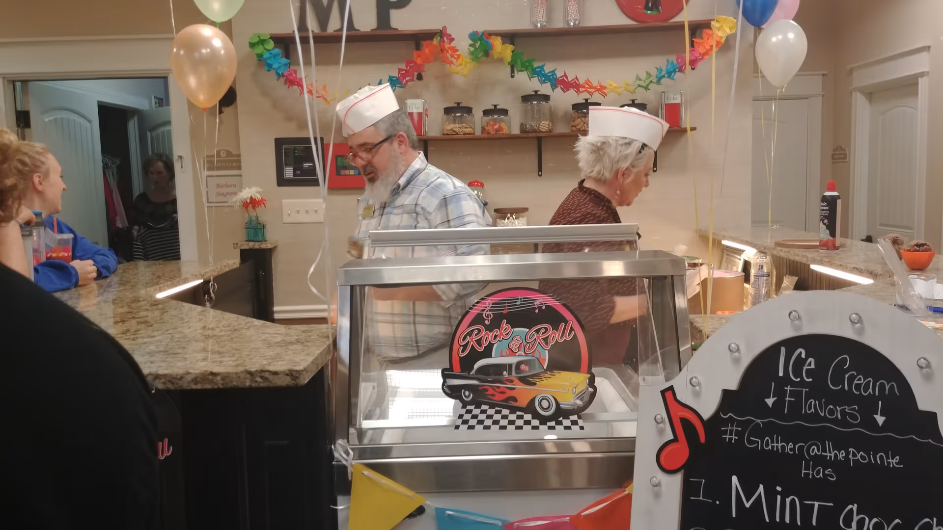 Two staff members wearing white paper hats stand behind a counter decorated with balloons and colorful paper garlands. In front of them is an ice cream display case with a 'Rock & Roll' sign featuring a classic car. A chalkboard sign nearby lists ice cream flavors, including Mint Chocolate. Several people are gathered around the counter in a festive atmosphere.