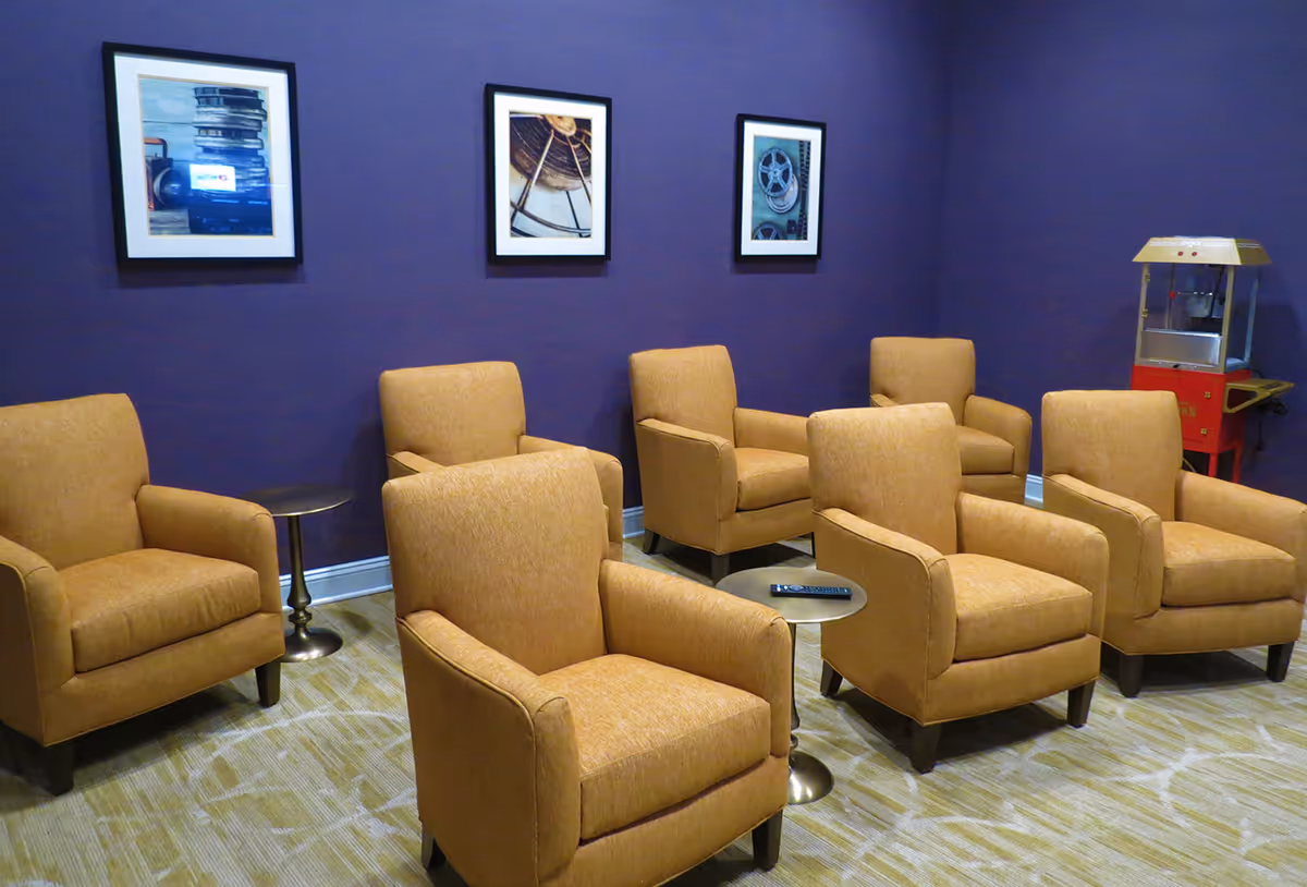 A small theater or media room with six tan upholstered armchairs arranged in two rows facing a screen or stage. The walls are painted dark purple and decorated with three framed pictures of film reels and cameras. A red and yellow popcorn machine is positioned in the corner of the room.