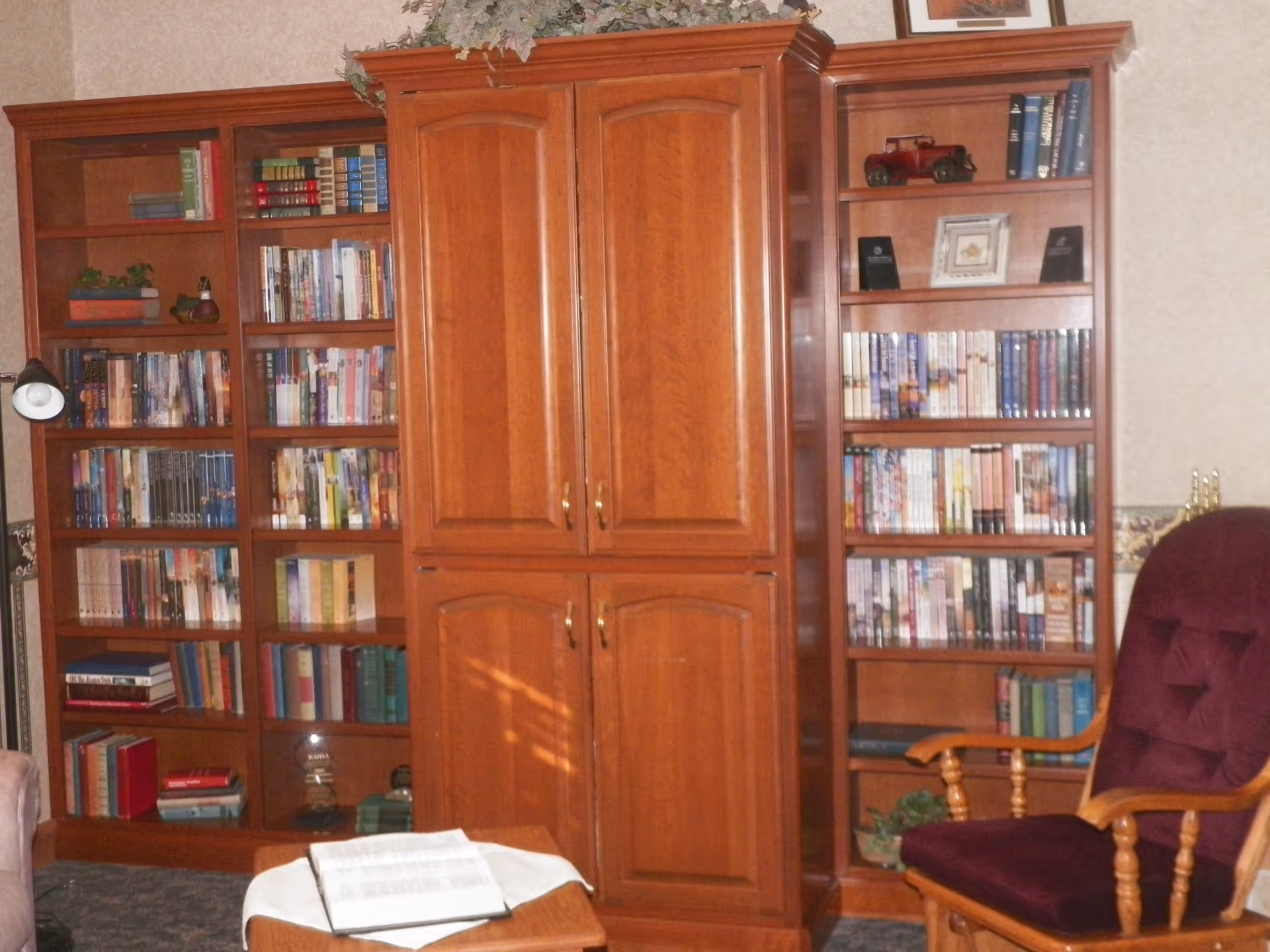 A cozy room with wooden bookshelves filled with books and decorative items, a wooden cabinet with closed doors in the center, a small wooden table with an open book on it, and a cushioned wooden armchair with a dark purple seat and backrest.