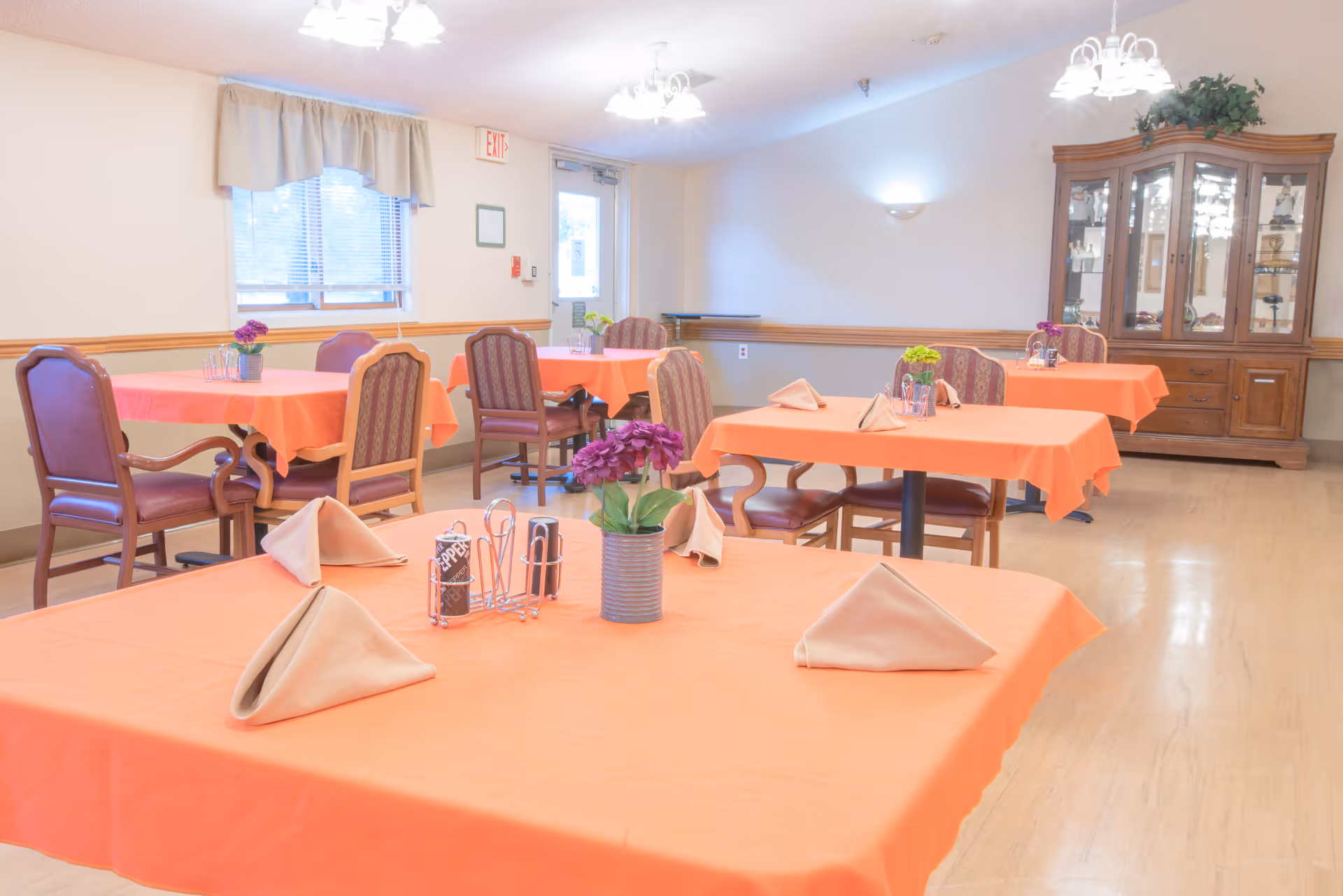 A dining room with several tables covered in orange tablecloths, each set with beige folded napkins, a small flower arrangement, and condiments. The room has wooden chairs with cushioned seats and backs, a wooden china cabinet with glass doors, and a window with beige curtains. The floor is light-colored wood, and the walls are painted white with wooden trim.