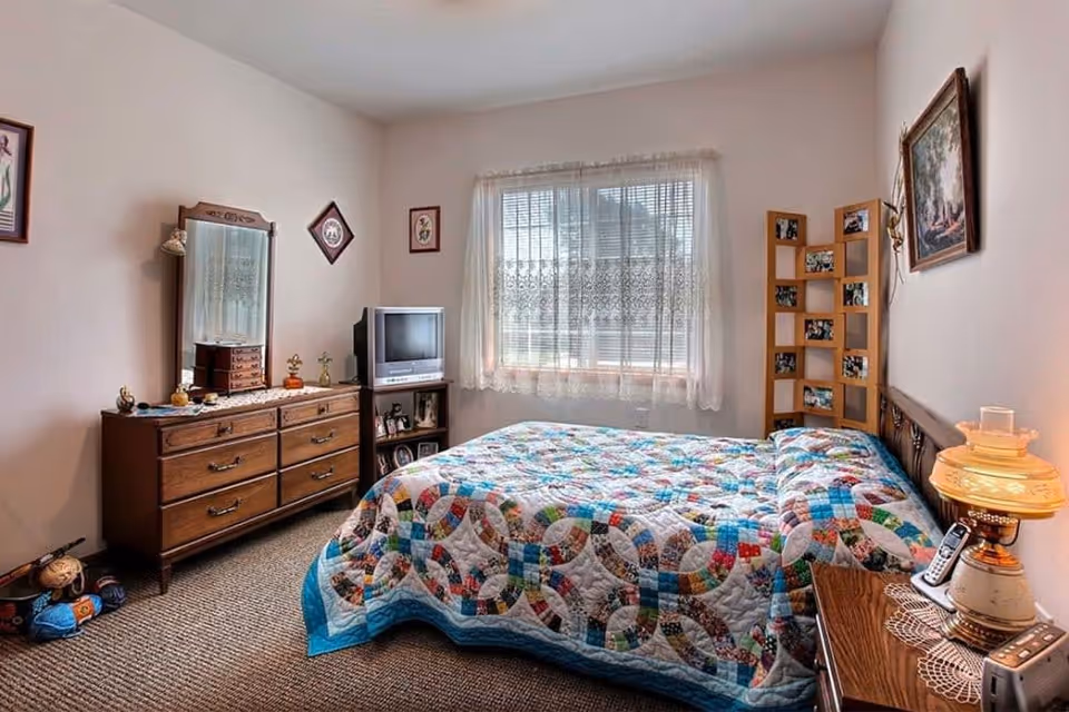 Cozy bedroom with a colorful patchwork quilt on the bed, wooden dresser with mirror, small TV, and a lace-curtained window.