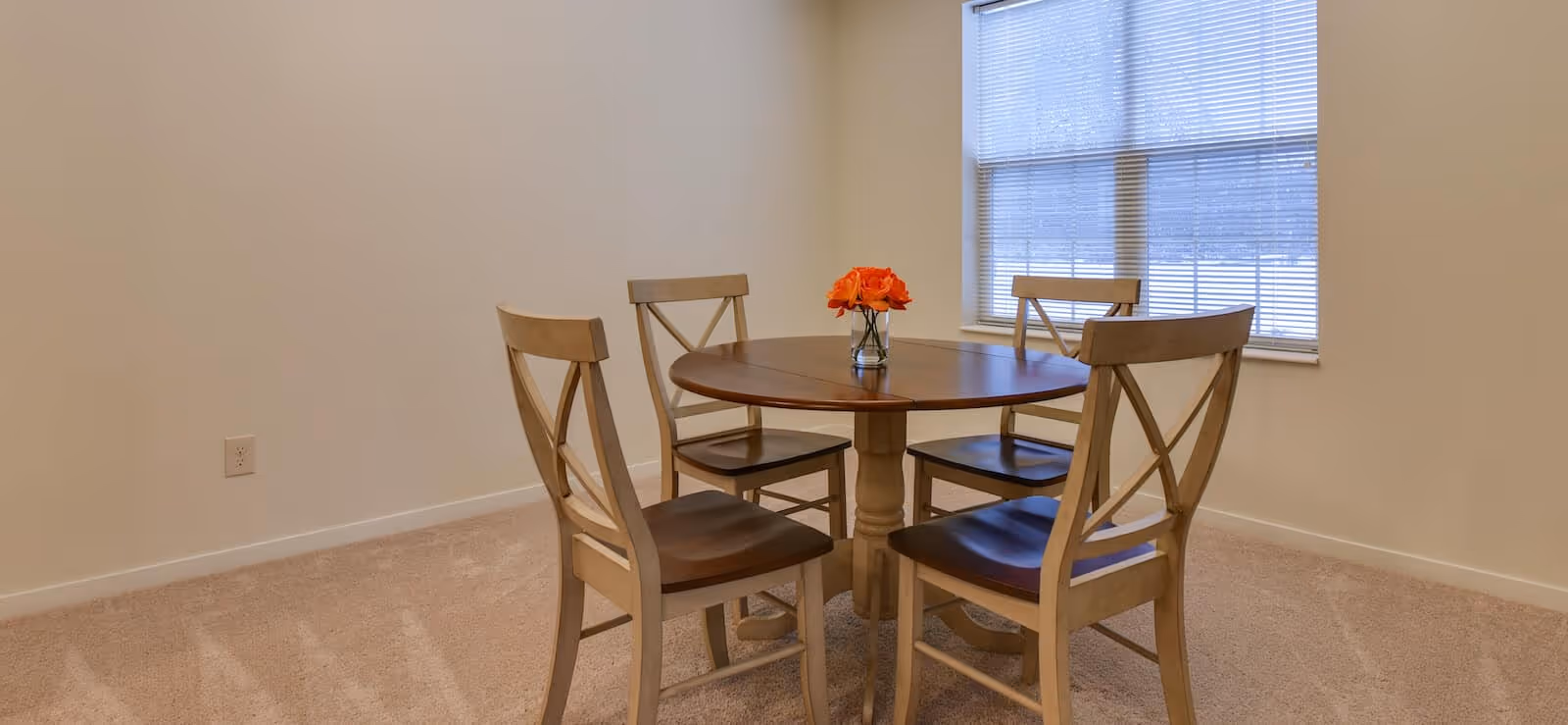 A simple dining area with a round wooden table and four matching wooden chairs with dark seats. A small vase with orange flowers is placed in the center of the table. The room has beige carpet and cream-colored walls, with a window covered by blinds letting in natural light.
