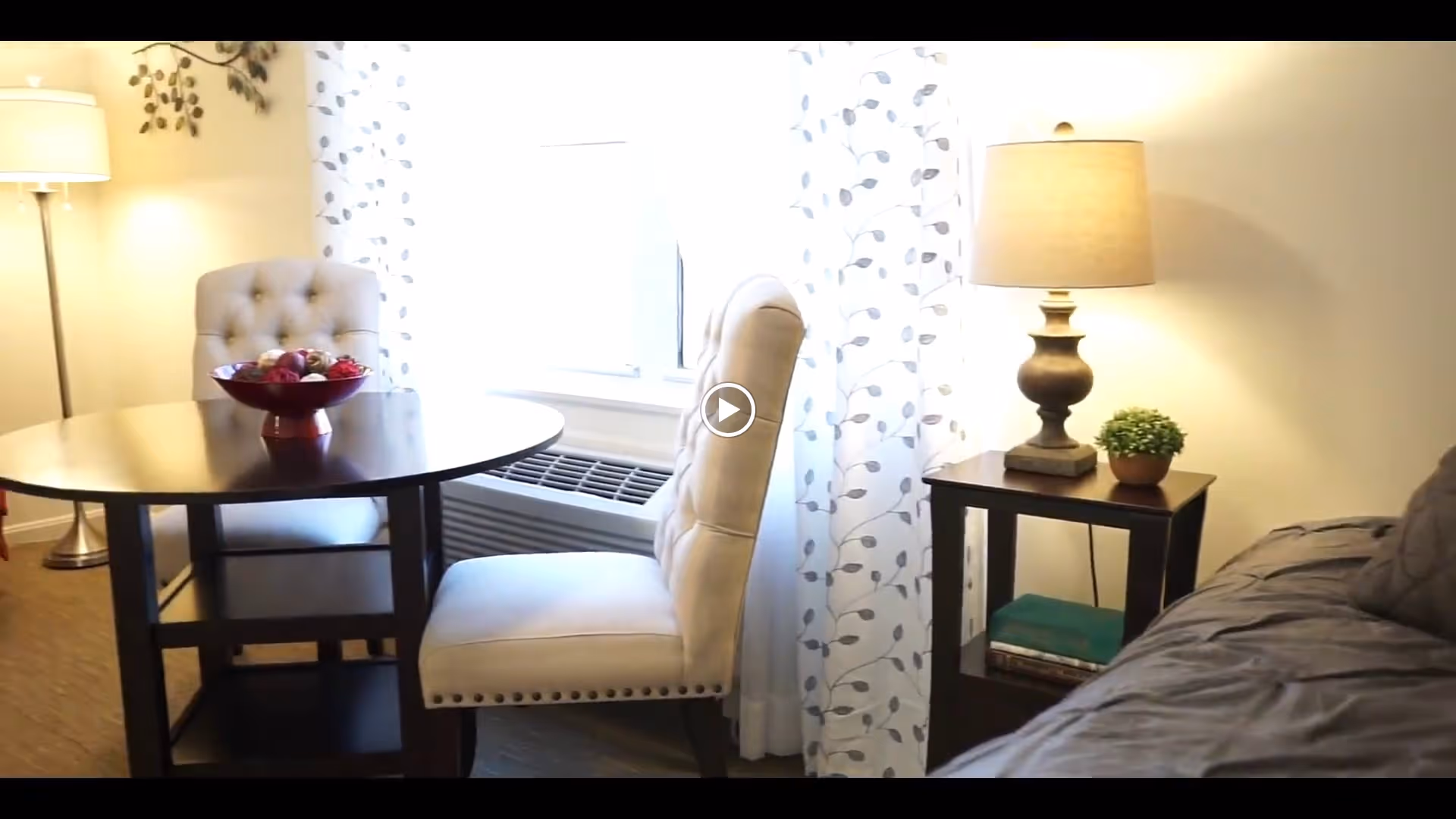 A cozy interior space featuring a round dark wooden table with a bowl of decorative items, two beige upholstered chairs, a window with patterned curtains, a floor lamp, and a side table with a lamp and a small potted plant next to a gray couch.