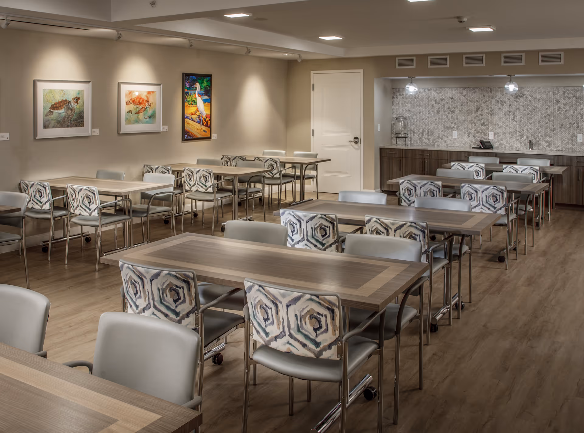 Communal dining room in a senior living facility with rows of wooden tables and patterned chairs, artwork on the walls, and a kitchenette along the back wall.