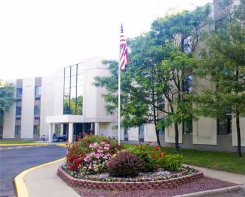 Front exterior view of a senior living facility with a flagpole, landscaped flower bed, and trees.