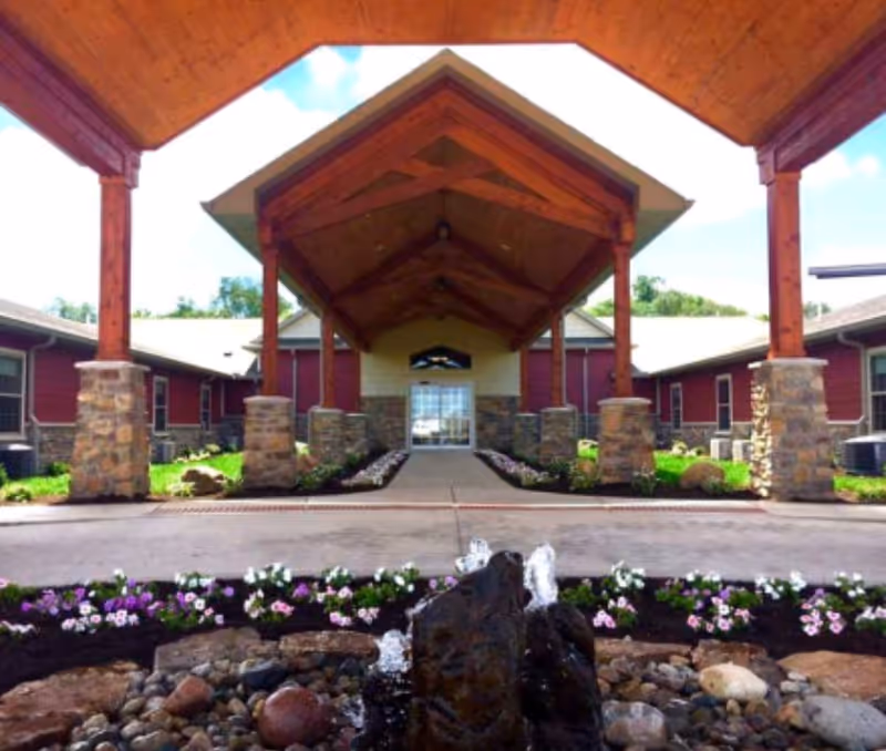 Entrance of Copper Creek Cottages Memory Care of Litchfield featuring a covered wooden canopy supported by stone pillars, a pathway lined with flowers, and a small water fountain with rocks in the foreground.