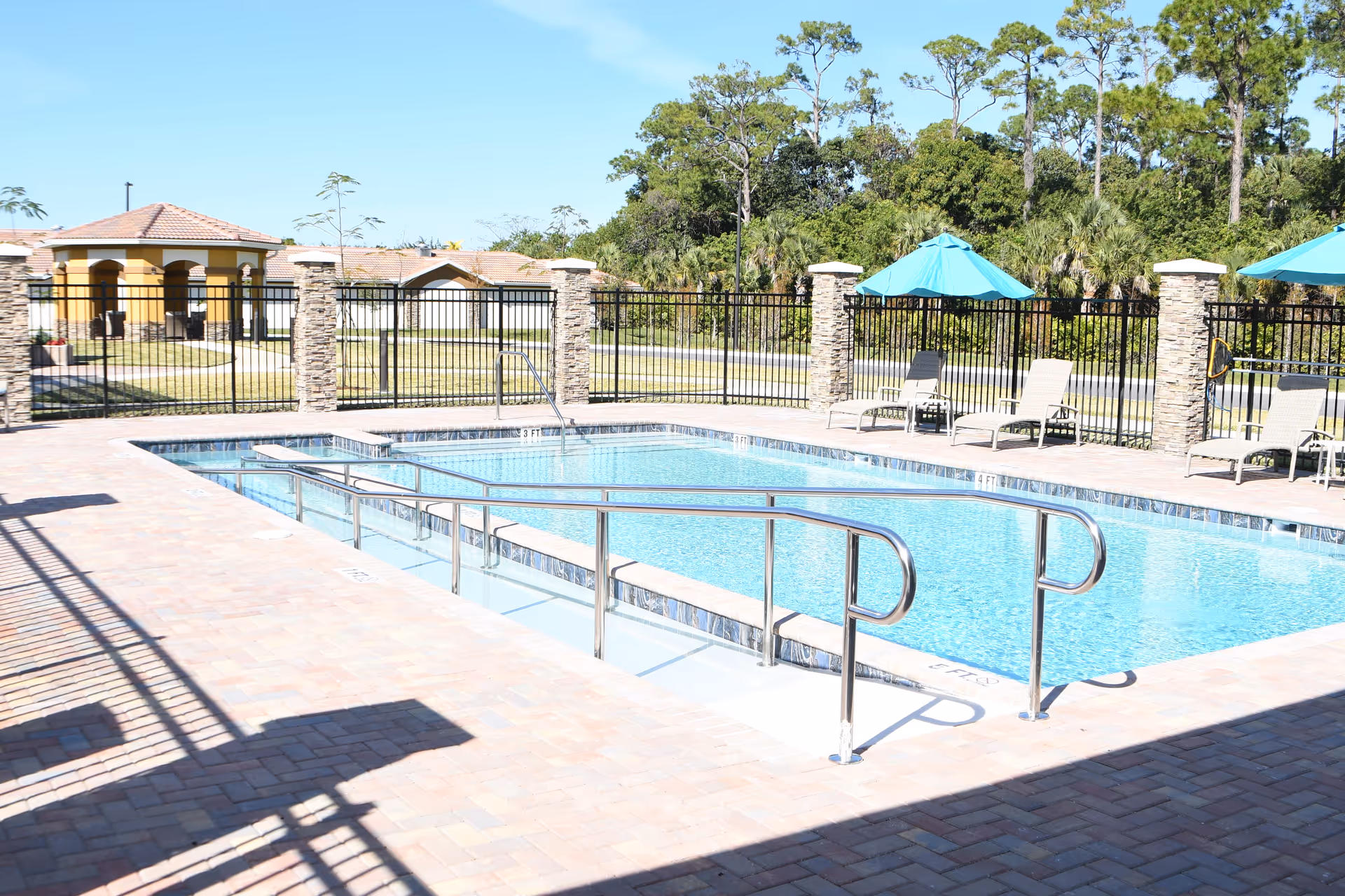 Outdoor swimming pool with metal handrails and a ramp for accessibility, surrounded by a brick patio. There are several lounge chairs with blue umbrellas along the fence, and trees and buildings are visible in the background under a clear blue sky.