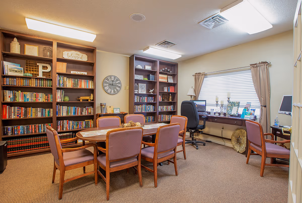 A senior living common room with a central table and chairs, bookshelves along the wall, and a desk with a computer by a window.